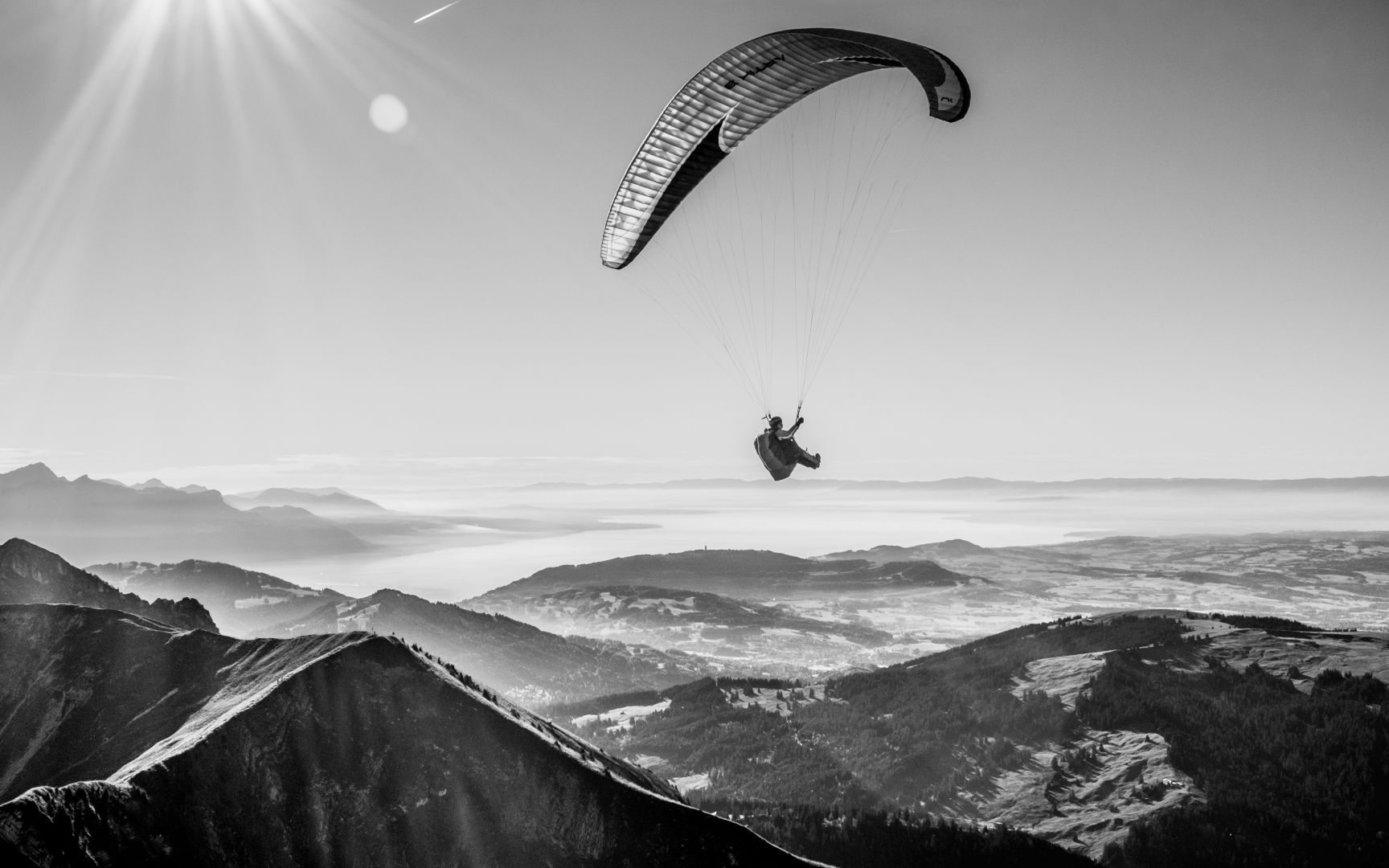 Un parapentiste dans les airs, volant au-dessus des montagnes qui se trouvent entre la Gruyère et la Riviera vaudoise (Montreux, Vevey, etc). Un peu de brume s’immisce entre les monts et tout au fond de la photo, on aperçoit le Lac Léman.