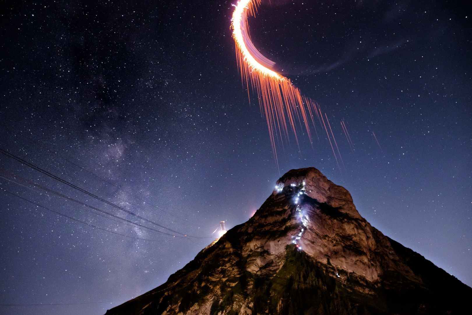 La montagne du Moléson la nuit. Une colonne de petites lumières serpente la montagne et se dirige vers le sommet. Un peu en contrebas vers la gauche, un point très lumineux indique que c’est l’arrivée du téléphérique. On peut d’ailleurs entre apercevoir les câbles, qui se détachent à peine du ciel étoilé, arriver vers ce point lumineux. Toujours vers la gauche de la photo, à côté de la montagne, on aperçoit la Voie lactée. Juste au-dessus du sommet de la montagne on voit un C lumineux avec des traînés se dirigeant vers le sommet.