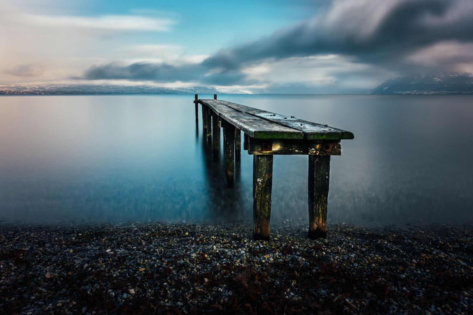 Un ponton en bois un peu vermoulu s’avance dans le lac depuis la plage de galets. Le ciel est changeant avec quelques endroits où on voit le ciel bleu, sinon ce sont des nuages gris foncés qui ont amené de la neige. Au loin, sur l’autre rive du lac, on aperçoit des montagnes enneigées. Les températures froides amènent une luminosité particulière dans le ciel et sur le lac entre le bleu de ciel et un blanc rosé. Le fait que la photo soit prise en pose longue fait que la surface de l’eau est bien lisse et que le déplacement des nuages causé par le vent d’hiver forment des sortes de rouleaux dans le ciel