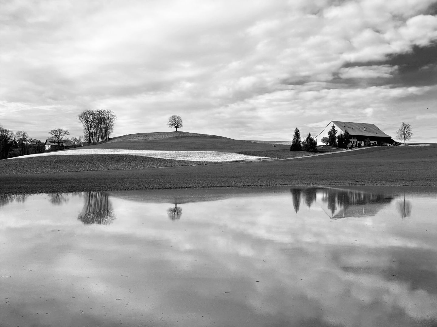 Image, en reflet dans l’eau d’un lac éphémère, d’une jolie ferme et de ses arbres qui l’entourent (sur la droite de la photo), d’un arbre solitaire au milieu et de quelques arbres et lointaines habitations sur la gauche de l’image. On peut voir aussi une petite bande de neige en dessous de l’arbre solitaire qui s’étend vers les groupe d’arbre sur la gauche.